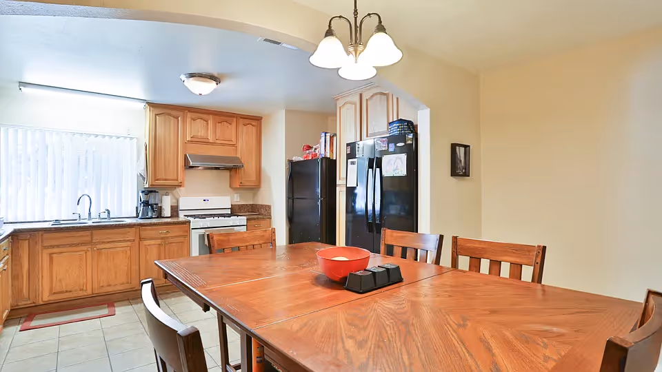 A kitchen and dining area with wooden cabinets, a white stove, a black refrigerator, and a wooden dining table with chairs. The kitchen has a window with vertical blinds and a sink with a faucet. There is a red bowl and a black condiment holder on the dining table.