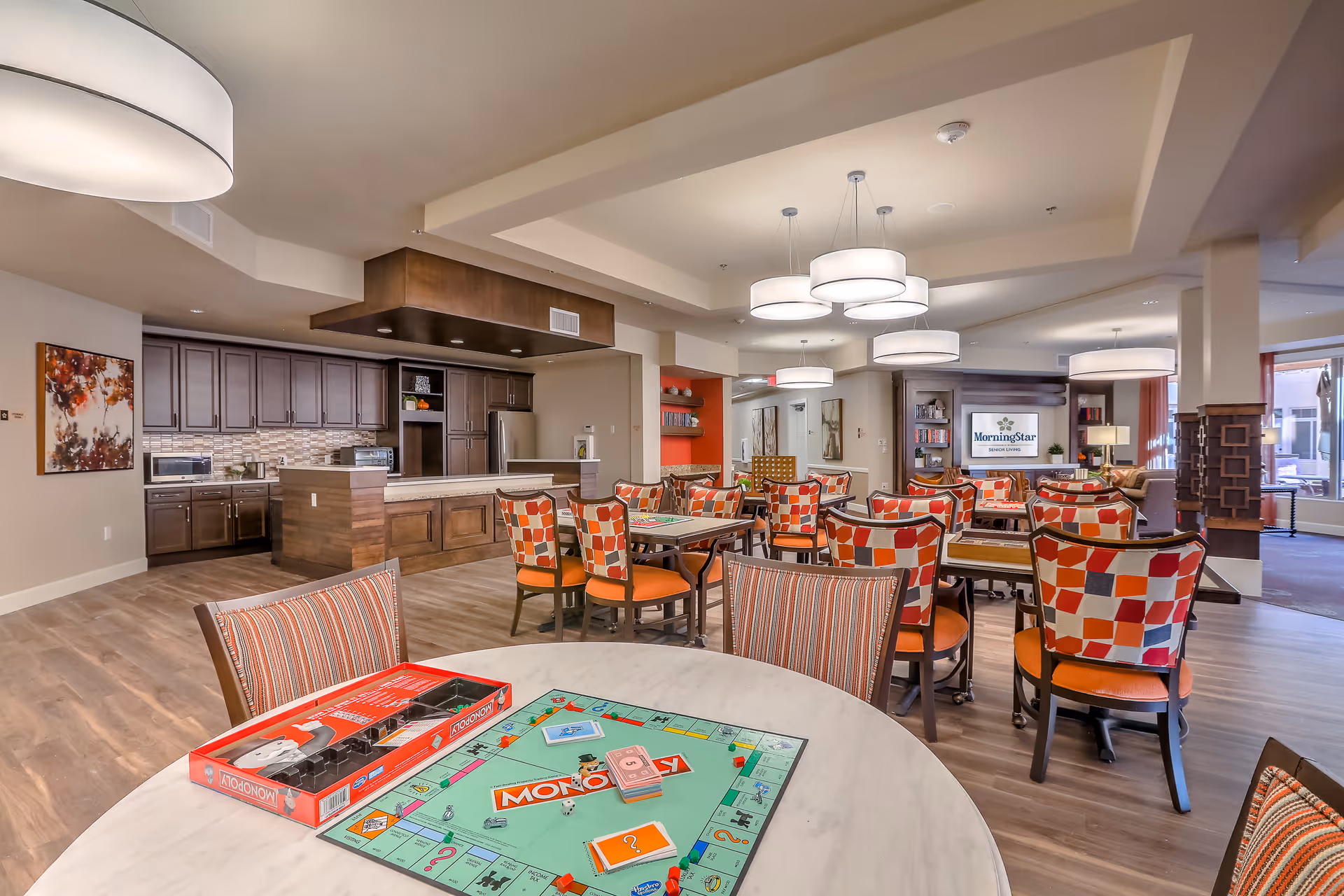 A spacious and well-lit common area in an assisted living facility featuring a game table with a Monopoly board in the foreground, multiple tables and chairs with colorful patterned upholstery, a kitchen area with dark wood cabinets and modern appliances, and a television displaying the MorningStar logo in the background.
