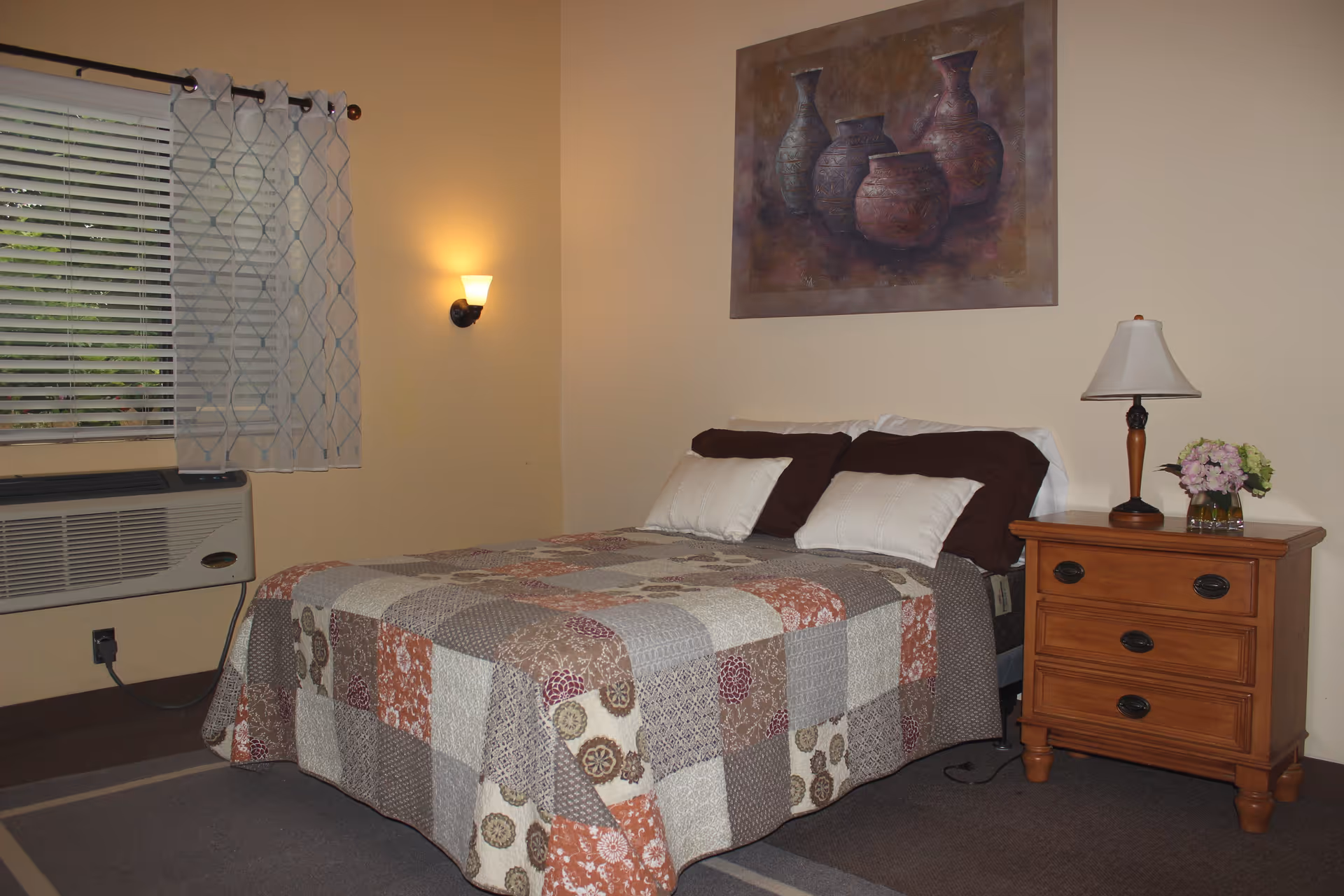 A cozy bedroom with a bed covered in a patchwork quilt featuring various patterns and colors. The bed has four pillows, two white and two dark brown. To the right of the bed is a wooden nightstand with three drawers, a table lamp, and a small vase with flowers. On the wall above the bed hangs a painting of four pottery vases. To the left, there is a window with blinds and a sheer curtain, and below it is an air conditioning unit. The walls are painted a light beige color, and a wall-mounted light fixture is on the wall near the window.