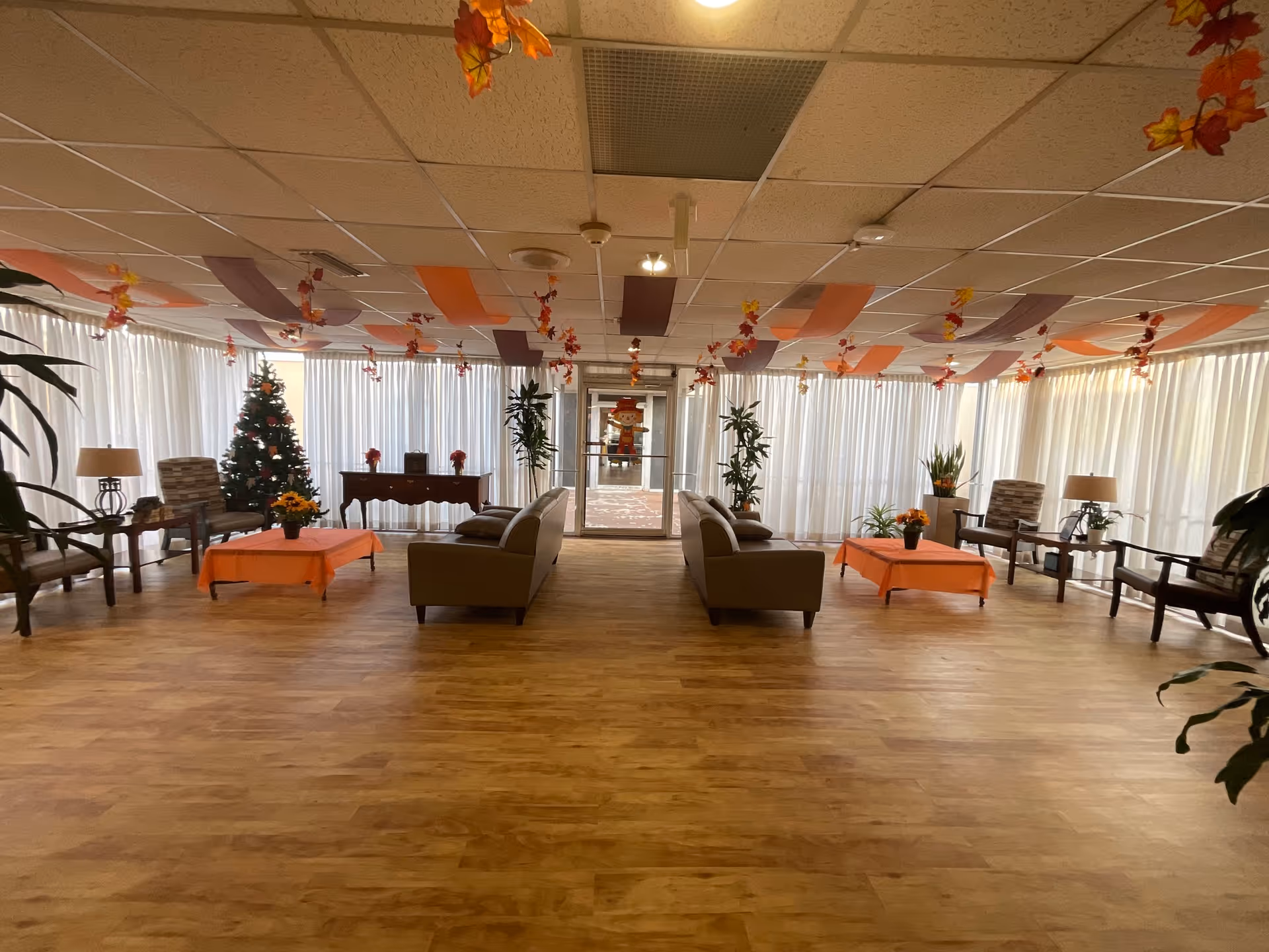Spacious common area with wooden flooring, two sofas facing each other with orange-covered coffee tables in between, several armchairs along the sides, potted plants, and a decorated Christmas tree near a window with white curtains. The ceiling is decorated with hanging orange, purple, and red streamers and autumn leaves. A glass door is visible at the far end of the room.