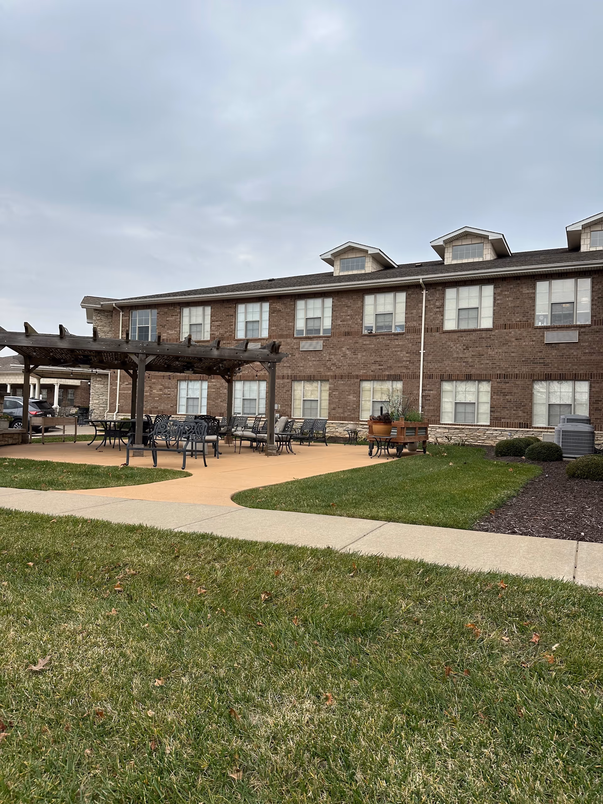 Outdoor patio with a pergola, tables and chairs in front of a two-story brick senior living building