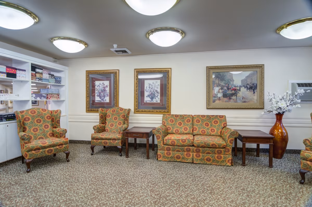 A senior-living common room with patterned armchairs and a matching sofa arranged with side tables, framed artwork on the wall, shelving, and a large decorative vase.