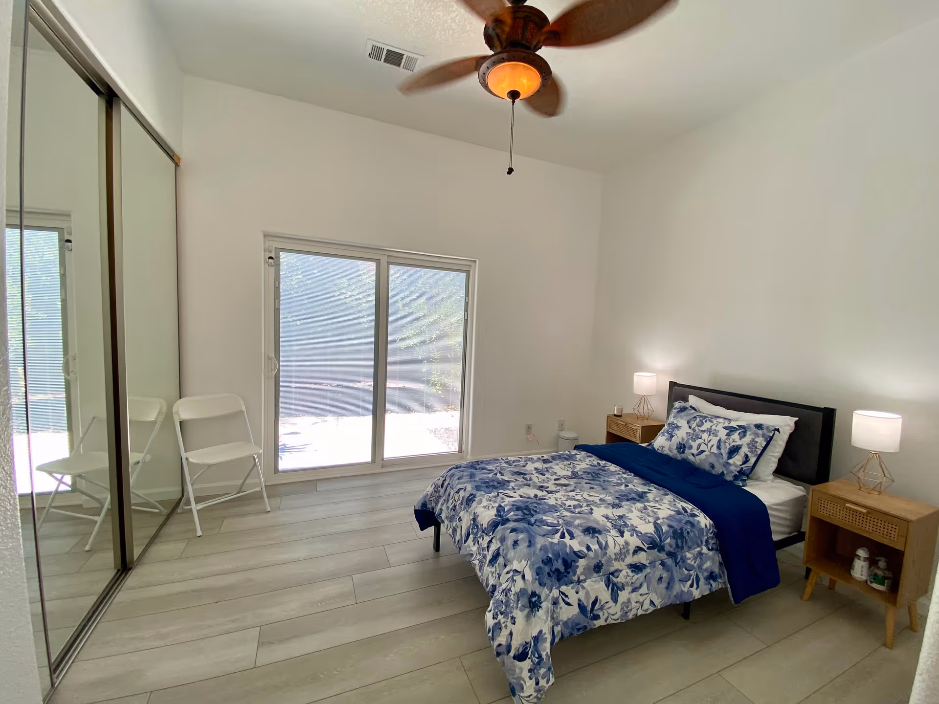 A bright bedroom with a single bed covered in blue and white floral bedding. There are two wooden nightstands on either side of the bed, each with a small lamp. A ceiling fan with a light is mounted on the white ceiling. A large sliding glass door with a view of greenery outside is on one wall, and a mirrored closet door is on the adjacent wall. A white folding chair is placed near the sliding door.