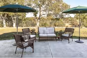 Outdoor patio area with a concrete floor featuring a cushioned loveseat, two armchairs, a small table, and two large green umbrellas providing shade. Trees and a grassy area are visible in the background.