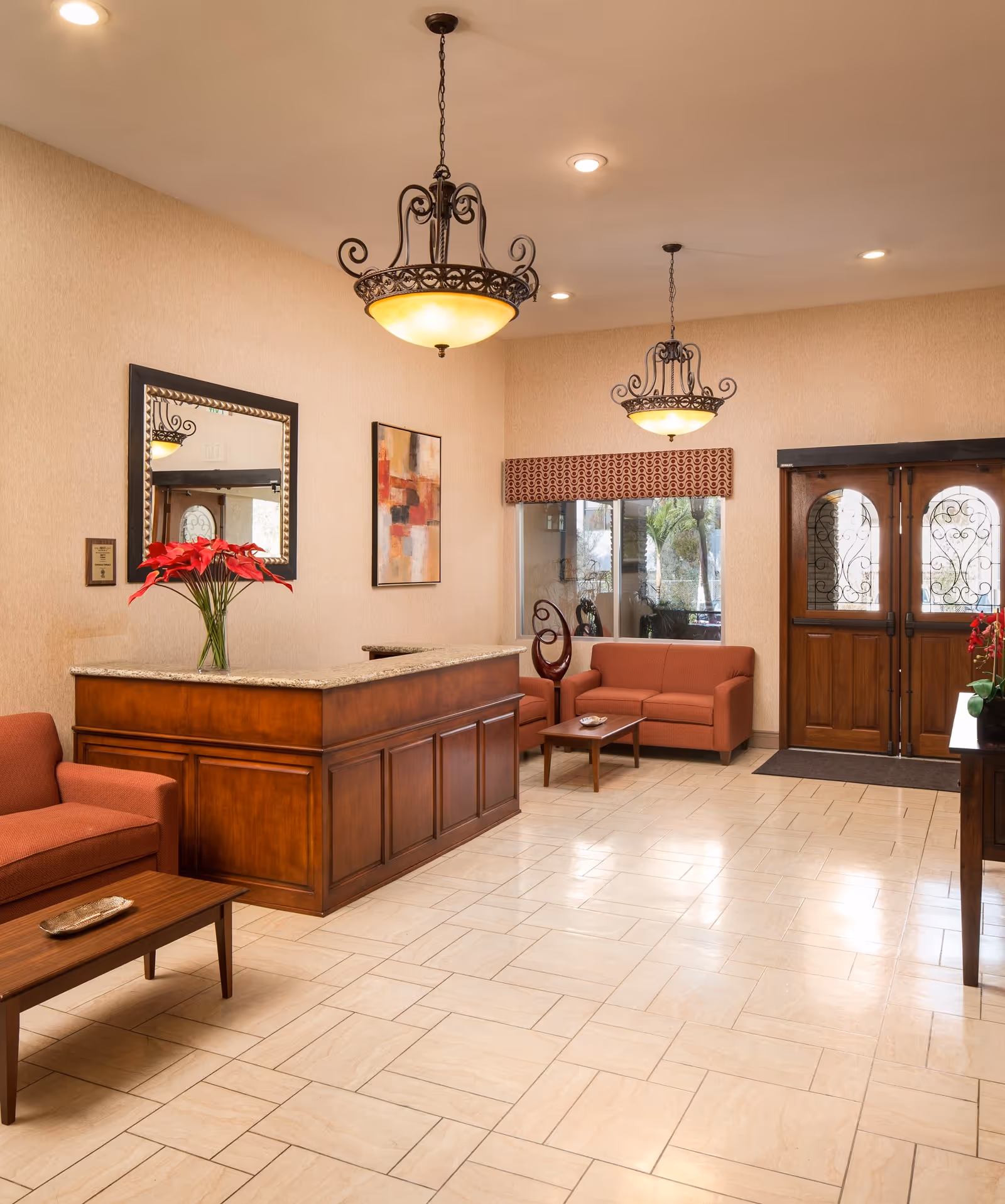 A warm and inviting reception area with a wooden front desk topped with a granite countertop. There are two red upholstered sofas and a matching armchair arranged around wooden coffee tables. The walls are beige with a large mirror and abstract artwork. Two ornate hanging light fixtures illuminate the space. Double wooden doors with decorative glass panels are visible in the background, along with a window with a patterned valance.