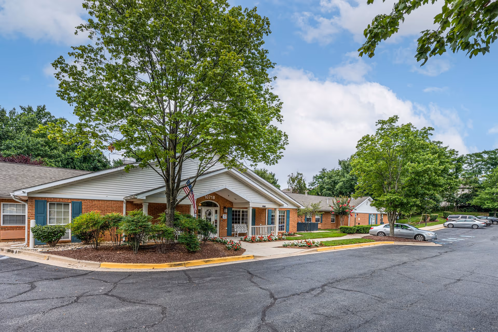 Exterior view of Arden Courts - ProMedica Memory Care Community in Potomac, showing a single-story brick building with white trim, a covered entrance with an American flag, surrounded by trees, shrubs, and a parking lot with several cars.