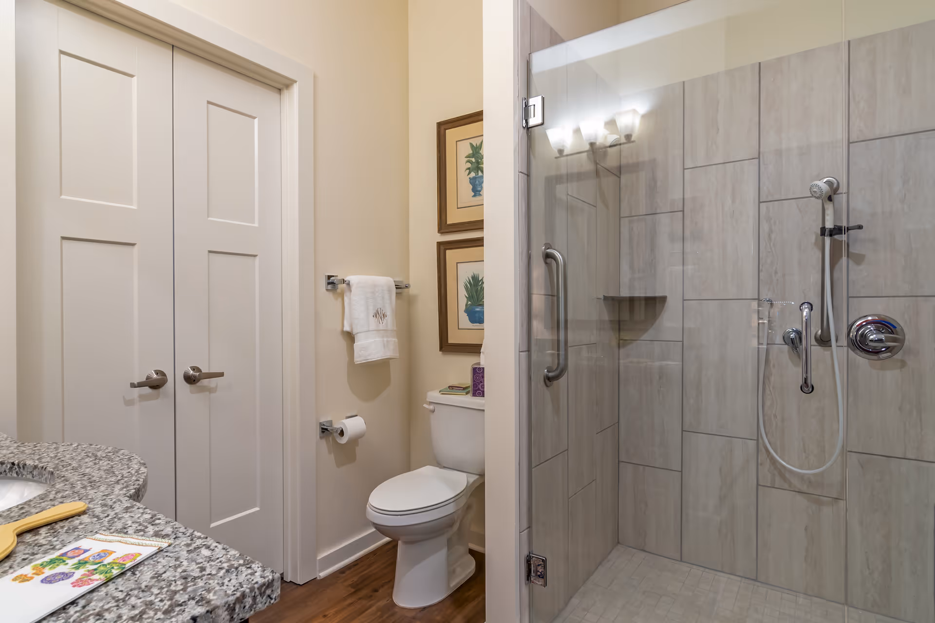 A clean and modern bathroom featuring a glass-enclosed shower with tiled walls and a handheld showerhead. Next to the shower is a white toilet with a towel rack above it holding a white towel. There are two framed botanical prints on the wall above the toilet. The bathroom has a granite countertop visible on the left side with a small mirror and some items on it. The floor is wooden, and there are double white doors in the background.