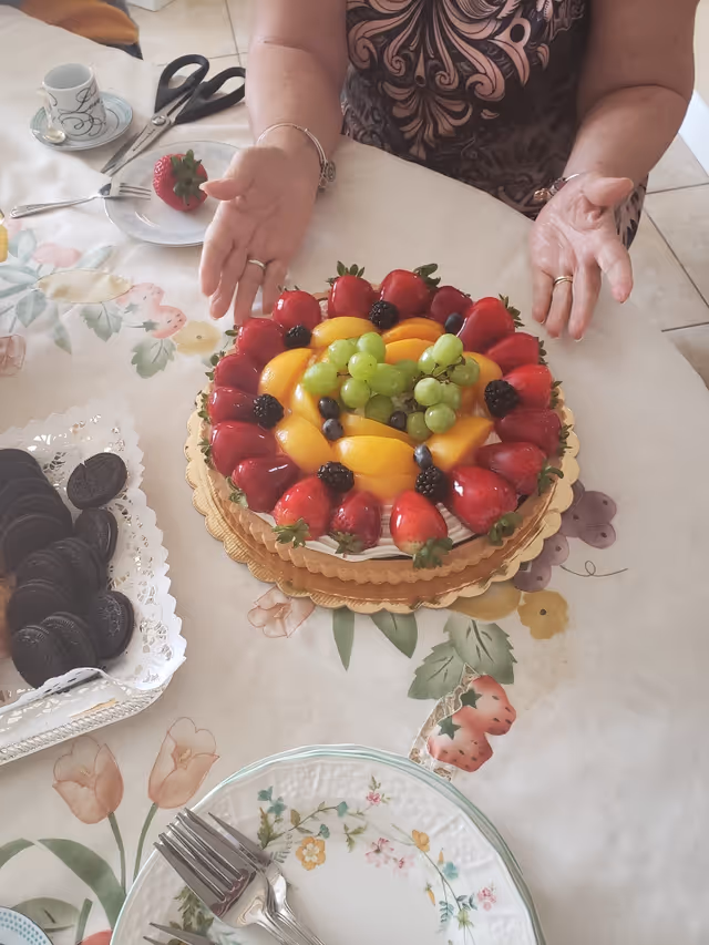 A fruit tart topped with strawberries, peaches, grapes and blackberries on a table while a woman gestures over it.