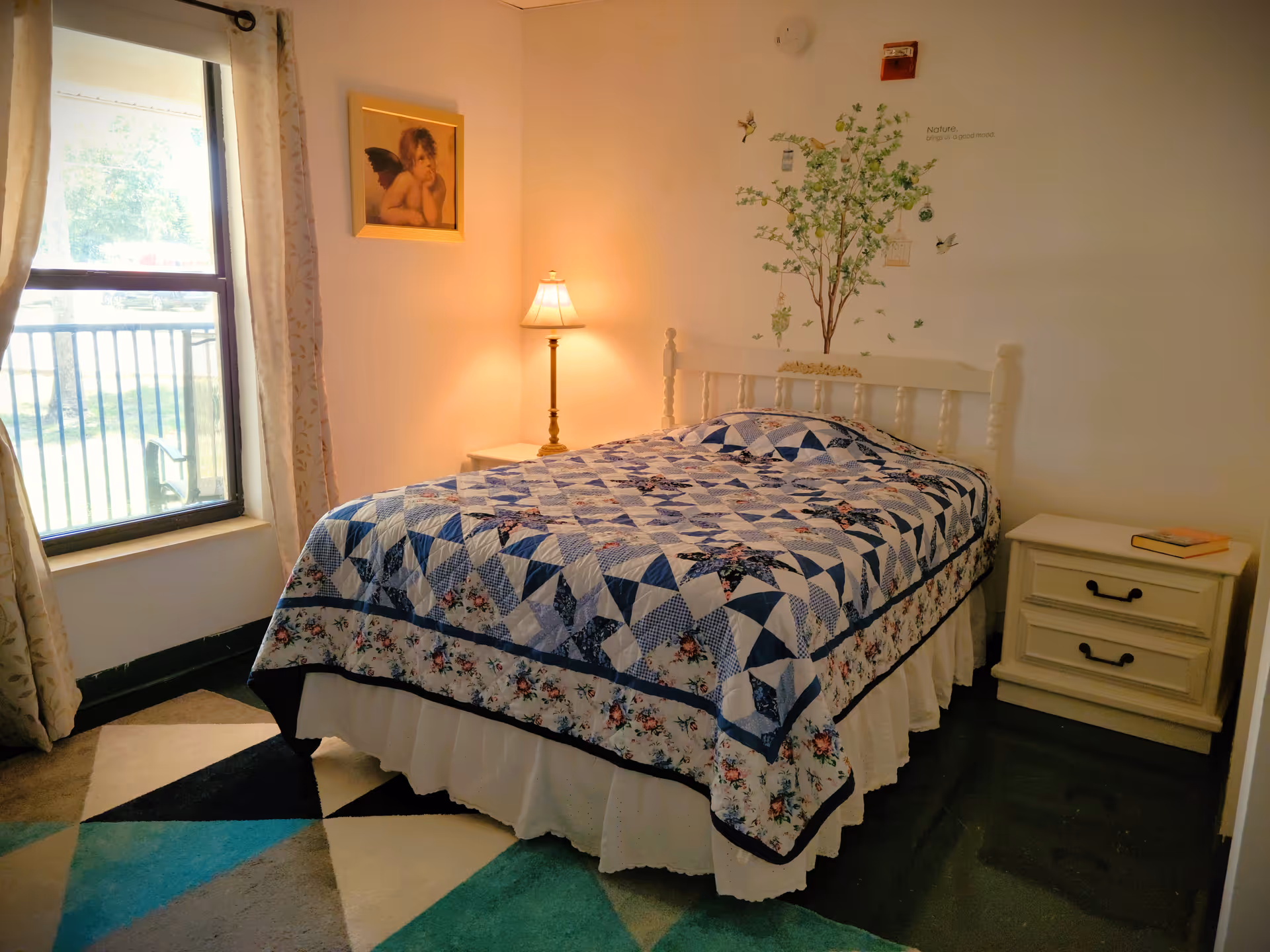 A cozy bedroom with a bed covered in a blue and white patterned quilt. There is a nightstand with a lamp and a book on either side of the bed. A window with curtains lets in natural light, and a framed picture of an angel hangs on the wall. A decorative tree decal with birds and the phrase 'Nature brings us a good mood' is on the wall above the bed.