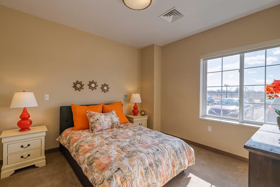 A cozy bedroom with a bed featuring a floral patterned comforter and orange pillows. There are two white nightstands on either side of the bed, each with a red lamp. Three decorative mirrors are hung on the beige wall above the bed. A large window on the right side lets in natural light and shows a parking lot outside. A wooden dresser with a vase of orange flowers is positioned under the window.