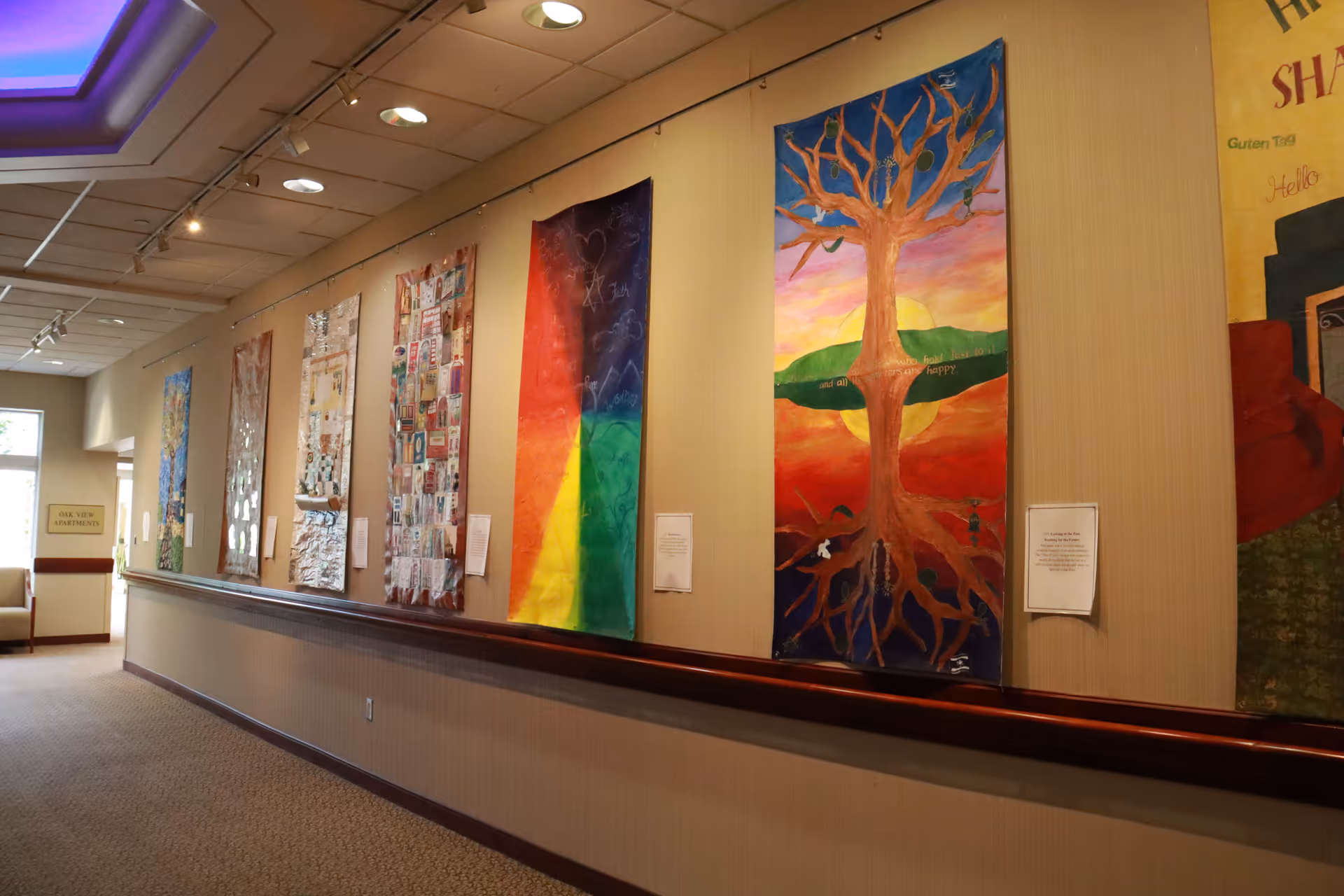 Interior hallway of a senior living facility with colorful artwork hanging on the wall. The hallway has beige walls, carpeted floor, and a wooden handrail running along the wall beneath the artwork. There is a sign at the end of the hallway indicating Oak View Apartments.