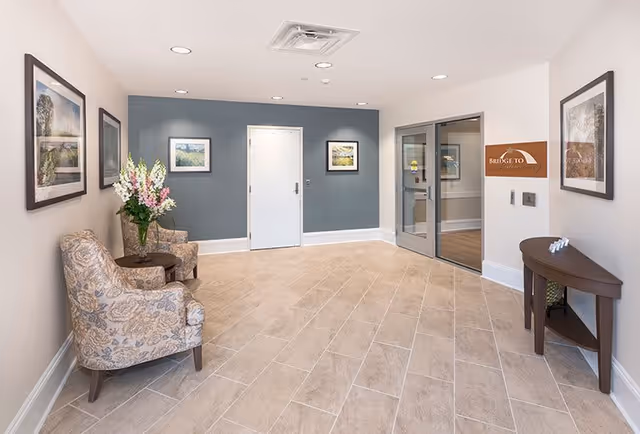 A clean and well-lit interior hallway area with beige tiled flooring and light-colored walls. There are two patterned armchairs with a small round table holding a vase of flowers on the left side. The back wall is painted blue-gray with a white door in the center and two framed pictures on either side. On the right side, there is a glass door next to a wooden console table with decorative items and a sign on the wall that reads 'Bridge To'. Several framed pictures hang on the walls.