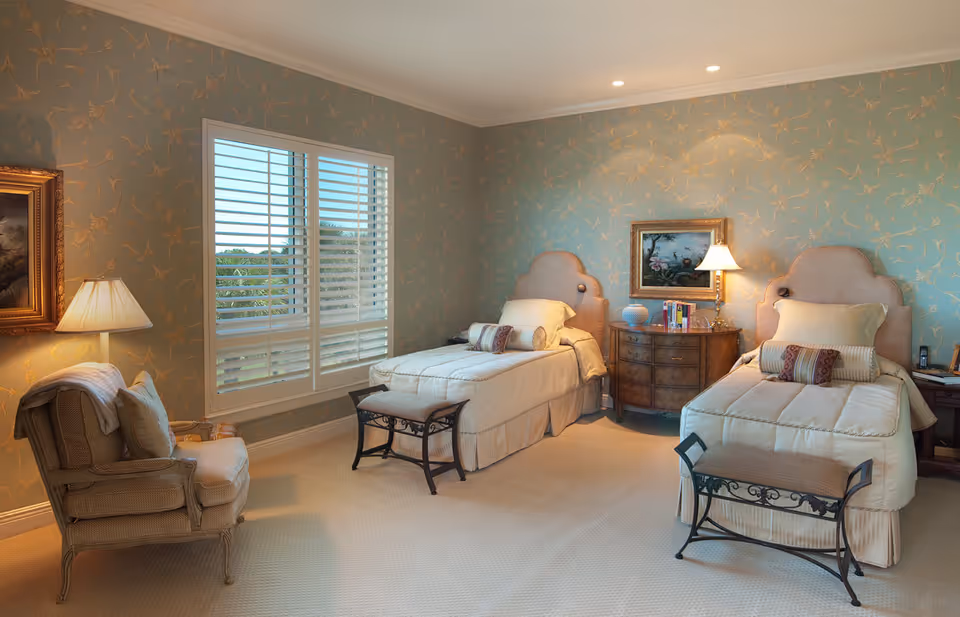 A cozy bedroom with two single beds, each with a padded headboard, beige bedding, and decorative pillows. Between the beds is a wooden nightstand with a lamp, books, and a decorative vase. A framed painting hangs above the nightstand. To the left, there is a cushioned armchair with a pillow and a blanket draped over the back. The walls are covered with light blue wallpaper featuring a subtle gold floral pattern, and a window with white plantation shutters lets in natural light.