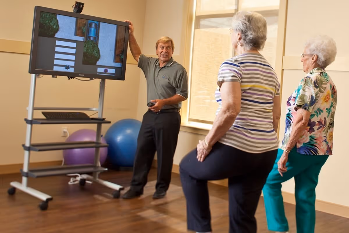 A man instructs two elderly women in a room with exercise balls and a large screen displaying a crosswalk and sidewalk scene. The women appear to be participating in a physical therapy or rehabilitation exercise.