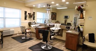 Interior view of a hair salon area within a senior living facility, featuring two salon chairs, a hair washing station, large mirrors, and various hair care products on the counter. The room has light-colored walls, wood flooring, and large windows allowing natural light.