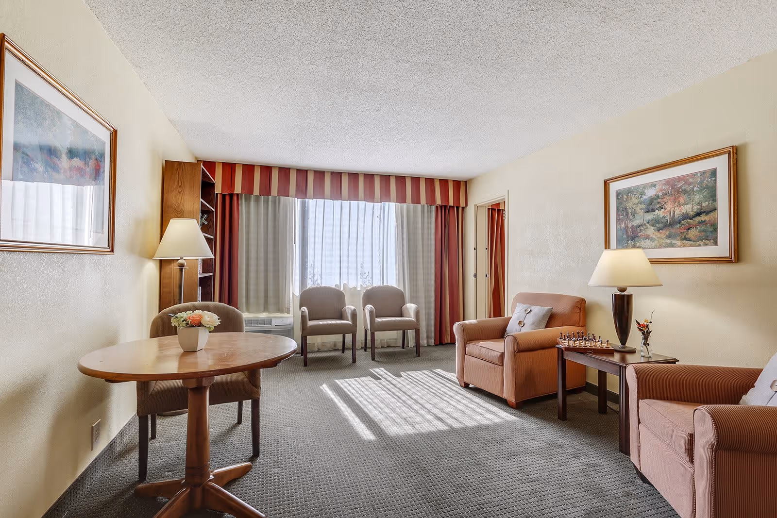 A bright senior living room with two armchairs, a small wooden table with a lamp and a chess set, two chairs near a window with striped curtains, a round wooden table with a flower pot, and framed paintings on the walls.