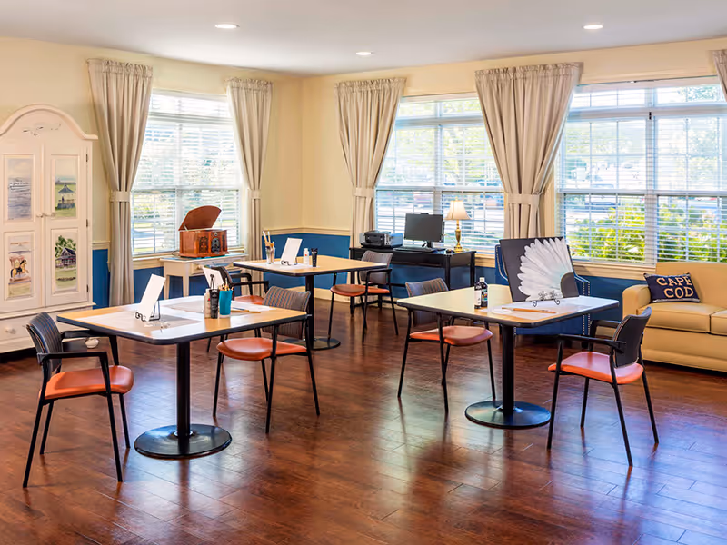 Bright communal room with several small tables and chairs, large windows with curtains, a couch and decorative cabinet.