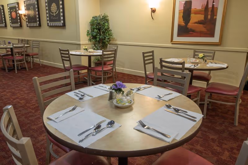 Dining area with round tables set with placemats, silverware, and small flower centerpieces. The room has red carpet flooring, beige walls with framed artwork, and several chairs with red cushions around the tables.