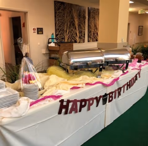A buffet table set up for a birthday celebration with chafing dishes, stacks of plates, and a 'HAPPY BIRTHDAY' banner hanging on the front. The table is covered with white and yellow cloths with pink accents. The background shows a hallway with framed artwork on the walls.