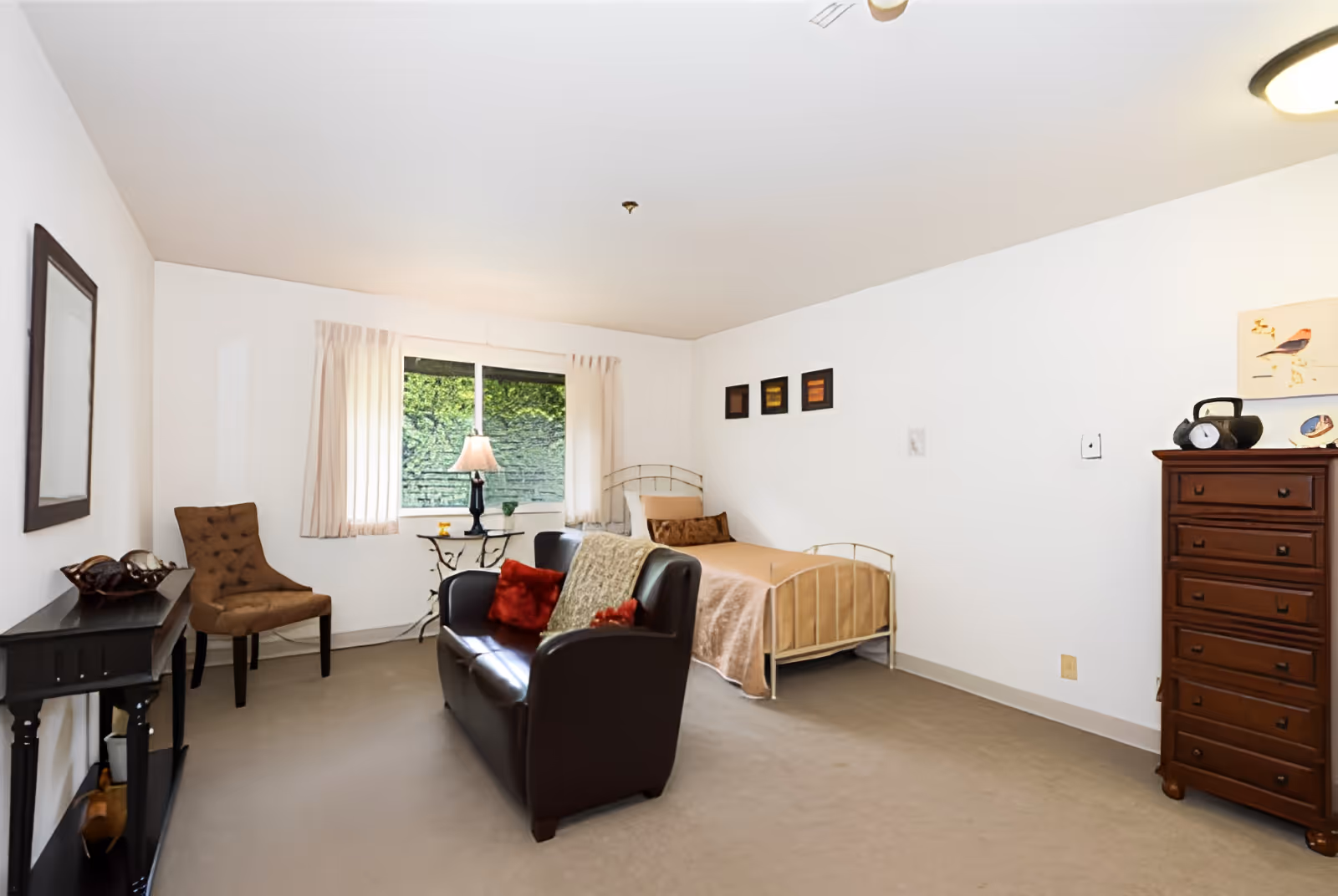 A cozy bedroom in a senior living facility featuring a single bed with beige bedding, a dark brown leather loveseat with red and beige pillows, a brown upholstered chair, a small black table with decorative items, a window with cream curtains, and a tall wooden dresser with a clock and artwork above it.