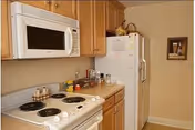 A small kitchen area with a white electric stove and oven, a white microwave mounted above the stove, wooden cabinets, a countertop with various items including a fruit basket and containers, and a white refrigerator. There is a framed picture on the wall next to the refrigerator.