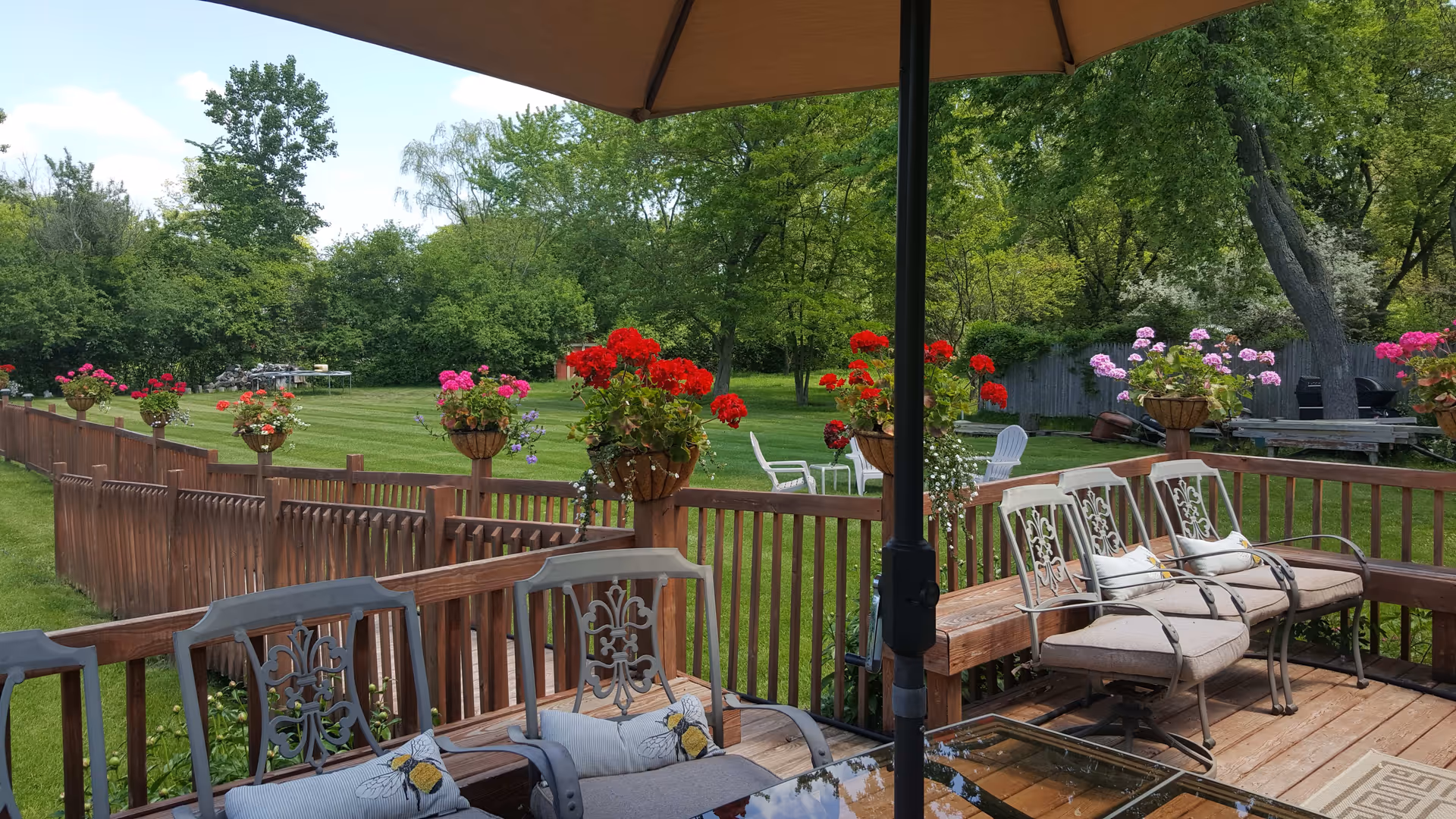 Outdoor wooden deck area with cushioned metal chairs and a glass-top table under a large umbrella. The deck overlooks a green lawn with a wooden fence adorned with hanging flower baskets containing red and pink flowers. Trees and shrubs are visible in the background under a partly cloudy sky.