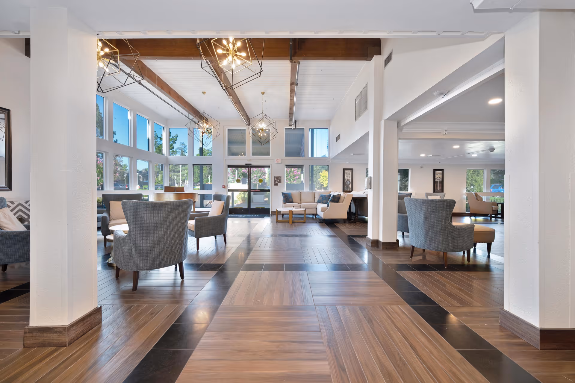 Spacious and well-lit senior living facility common area with large windows, modern pendant lights, wooden beams on the ceiling, and comfortable seating including armchairs and sofas arranged in small groups.