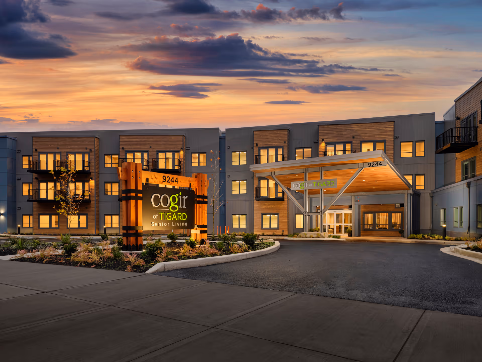 Exterior view of Cogir of Tigard senior living facility at sunset, showing a modern three-story building with balconies, illuminated windows, a large wooden sign with the facility name, and a covered entrance.
