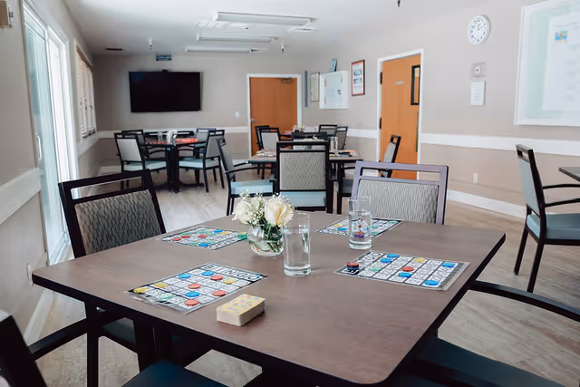 A well-lit dining or activity room with several tables and chairs. On the closest table, there are bingo cards, colorful bingo chips, a deck of cards, two glasses of water, and a small vase with white flowers. The room has light-colored walls, a wall-mounted TV, and wooden doors in the background.