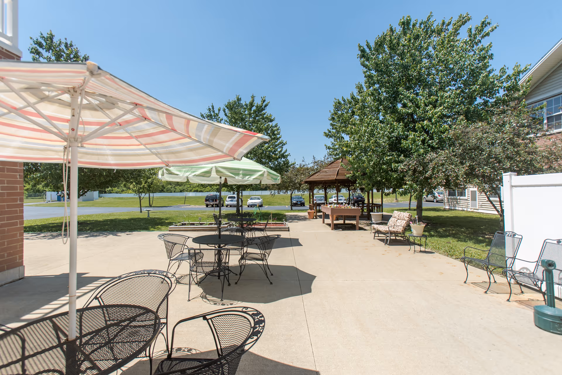 Outdoor patio area with metal tables and chairs under large umbrellas, surrounded by trees and greenery. There is a gazebo in the background and a few parked cars visible beyond the grassy area.