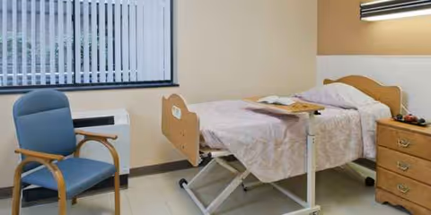 A senior living patient room featuring a single bed, blue chair, and wooden dresser.