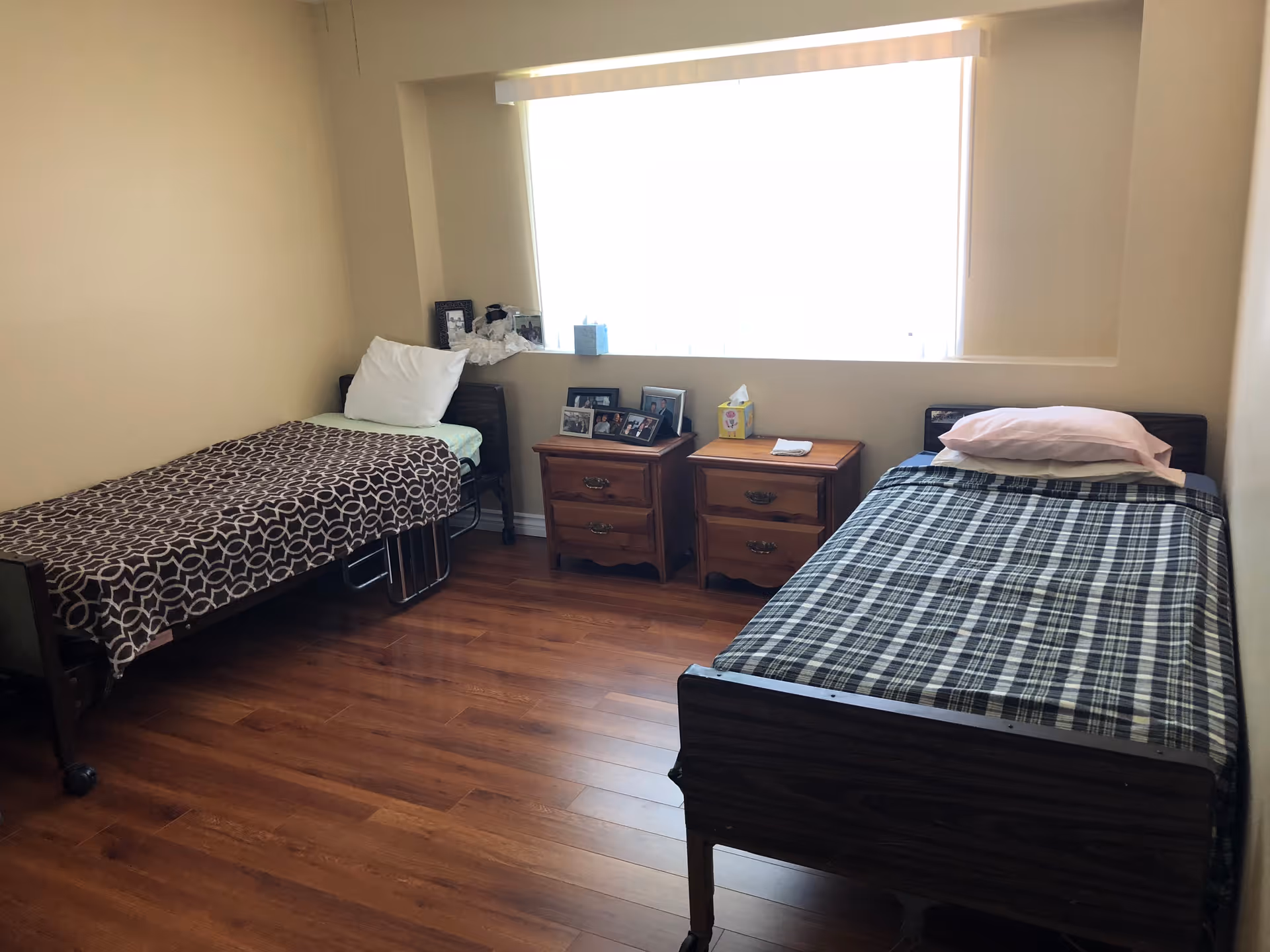 Bright shared bedroom with two single beds, wooden nightstands holding framed photos, and a large window.
