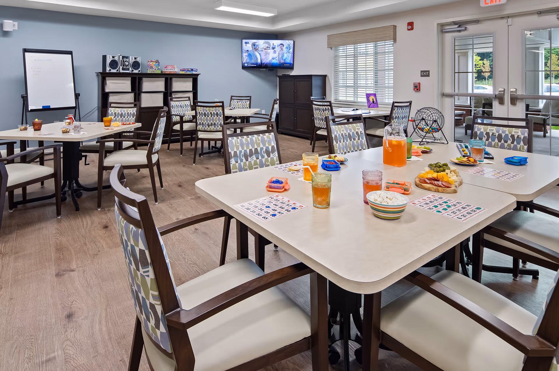 A bright and spacious activity room with several tables and chairs arranged for a group game of bingo. The tables have bingo cards, snacks, drinks, and a bingo cage with balls. There is a TV mounted on the wall, a whiteboard, and shelves with board games and a stereo system. Large windows and glass doors let in natural light and provide a view of the outdoor area.