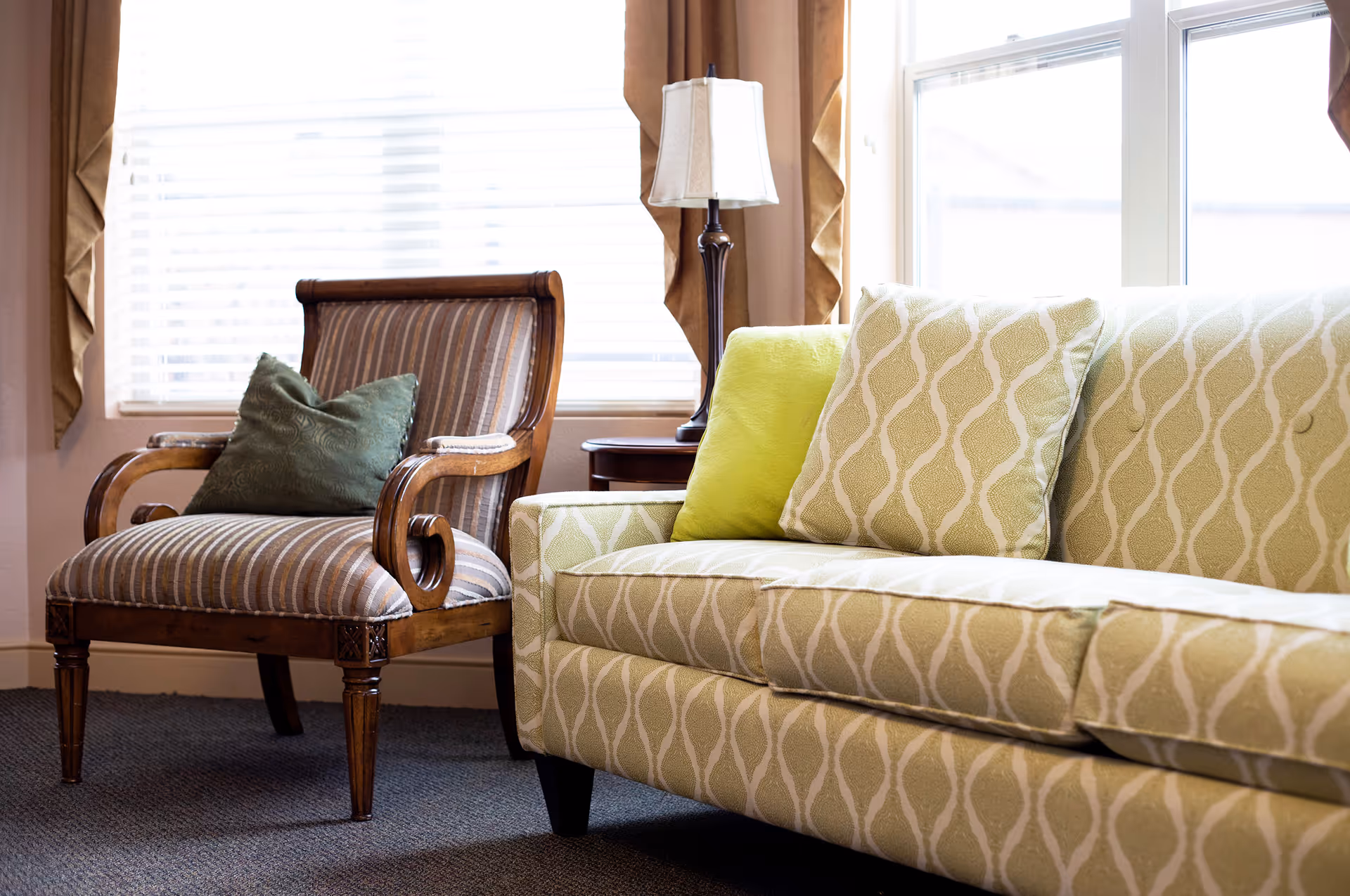 Sunlit sitting area with a patterned sofa, a striped wooden armchair with a pillow, and a floor lamp by a window.