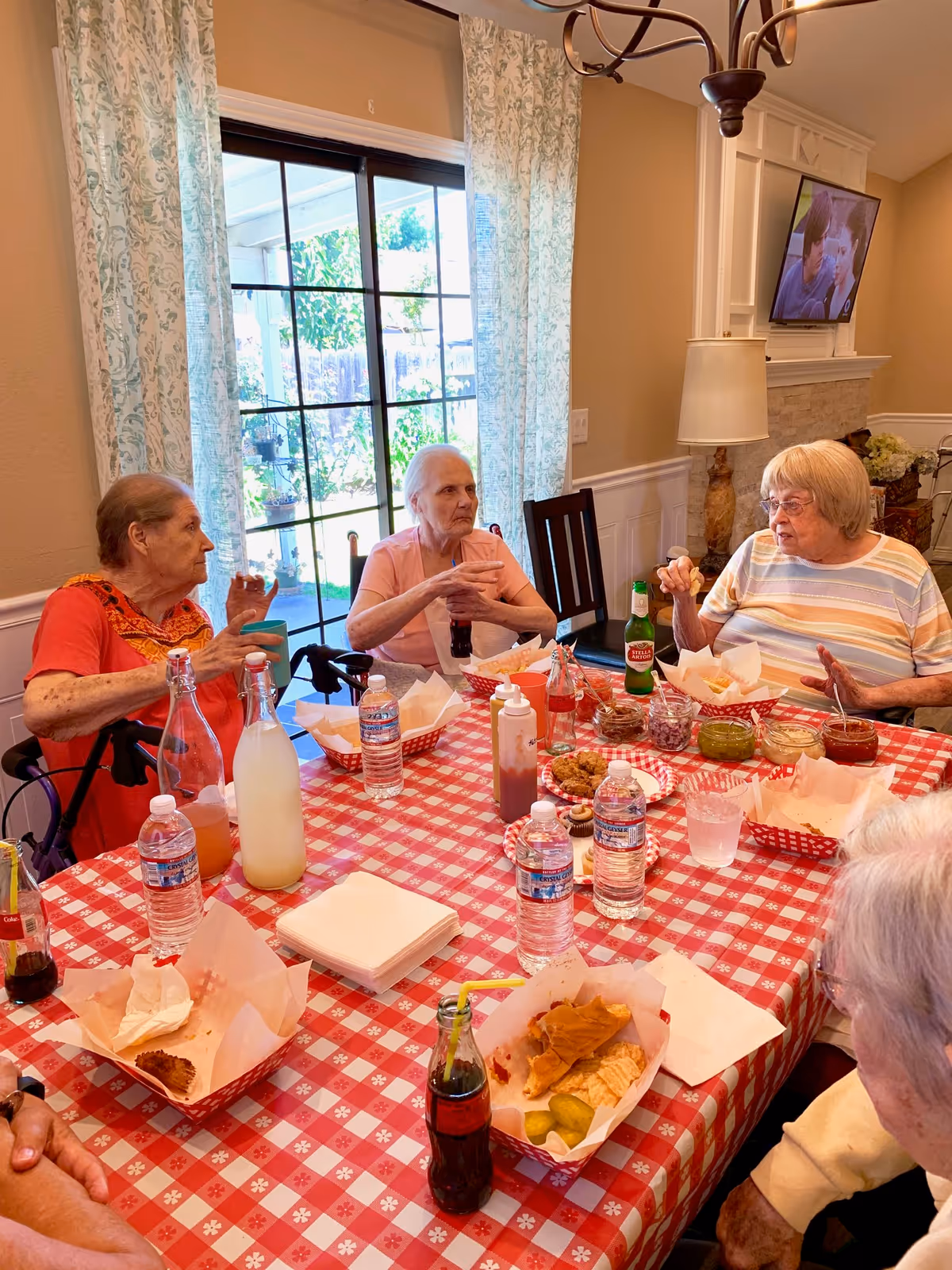 Four elderly women sitting around a dining table with a red and white checkered tablecloth, eating and drinking. The table has various food items, condiments, bottled water, and soda. The room has beige walls, a large window with patterned curtains, a lamp, and a TV mounted on the wall.