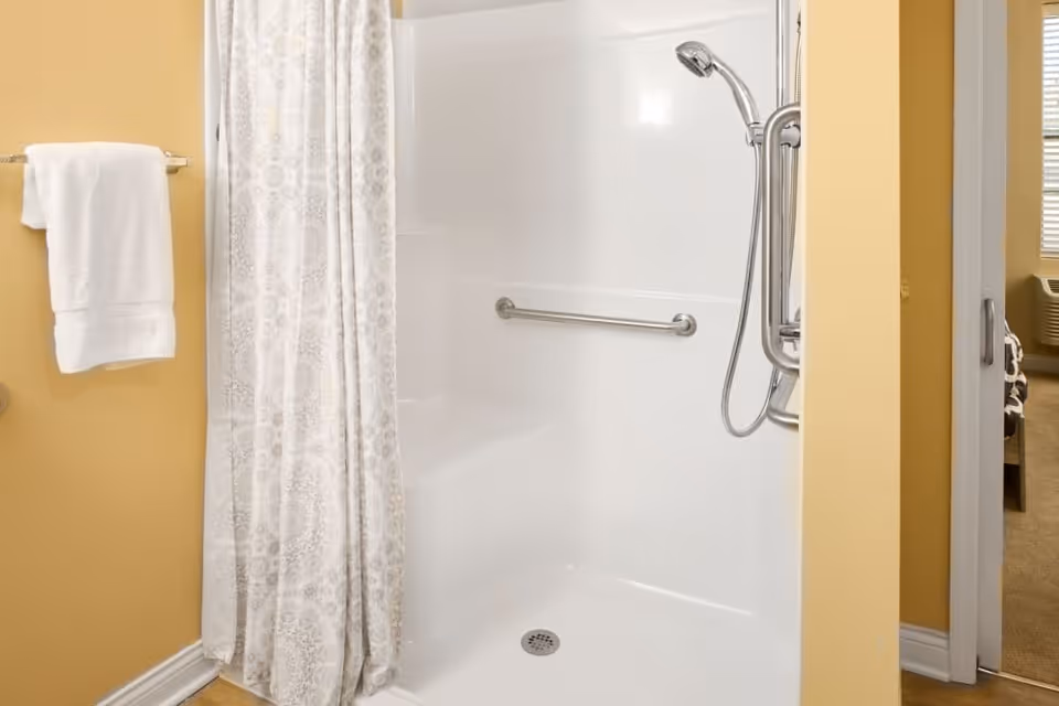A clean, accessible shower area with a white shower curtain, a grab bar on the wall, and a handheld showerhead. A white towel hangs on a towel rack mounted on a yellow wall next to the shower. Part of an adjacent room with carpet and a window air conditioner is visible through an open doorway.
