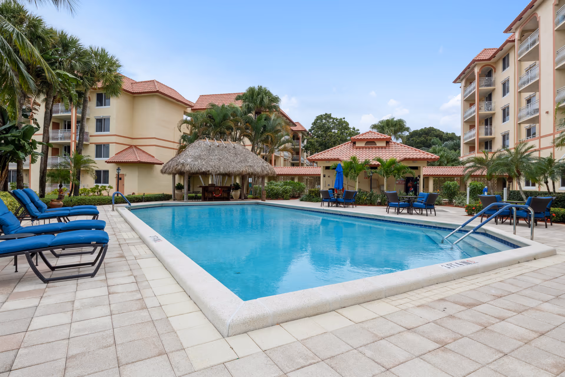 Outdoor swimming pool area at The Horizon Club with blue lounge chairs on the left, a thatched-roof gazebo, palm trees, and multi-story residential buildings in the background under a partly cloudy sky.
