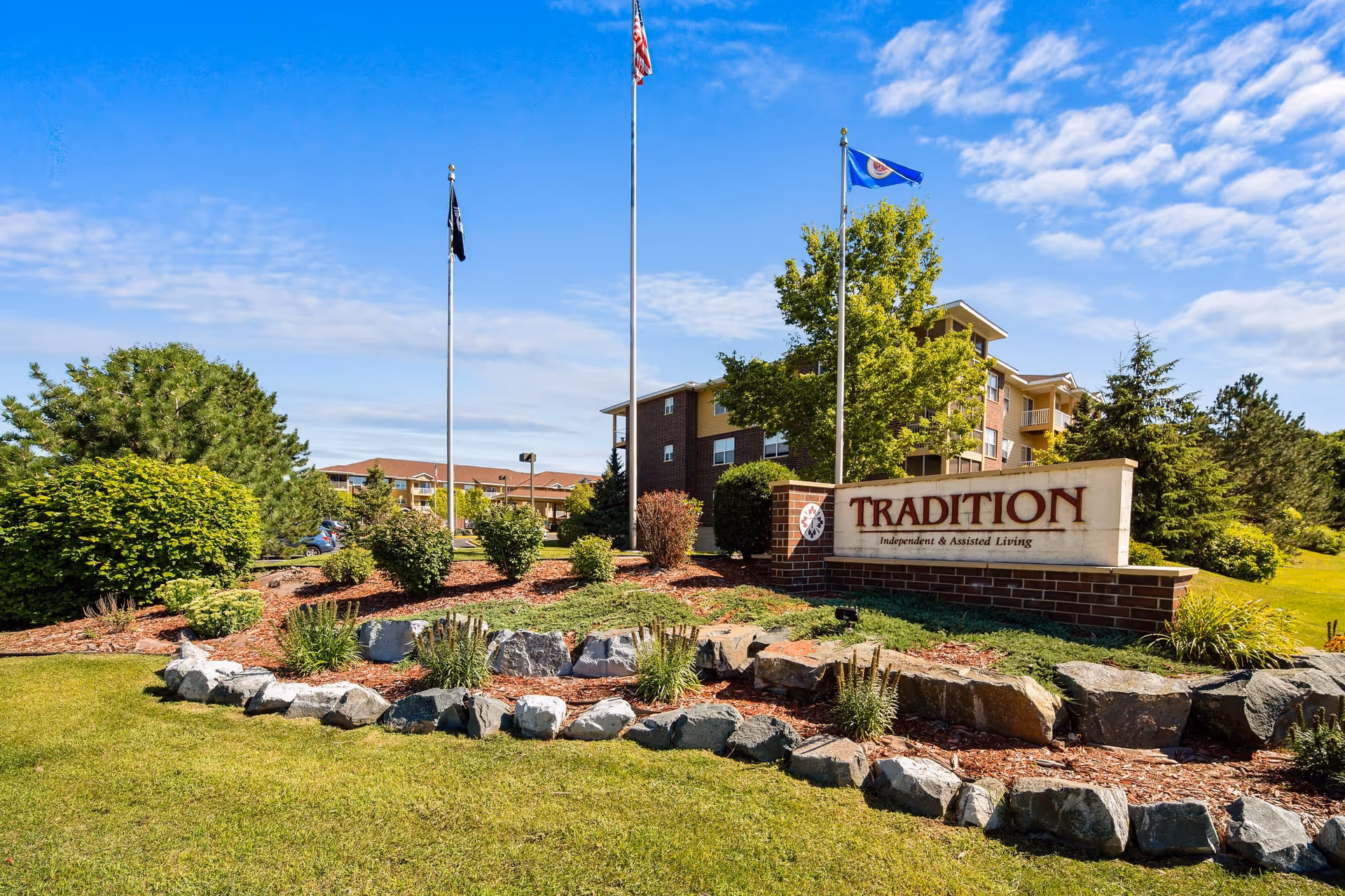 Outdoor view of Tradition Living facility with a large sign reading 'TRADITION Independent & Assisted Living' surrounded by landscaped bushes, rocks, and grass. Three flagpoles with flags are visible behind the sign, and a multi-story building is in the background under a partly cloudy blue sky.
