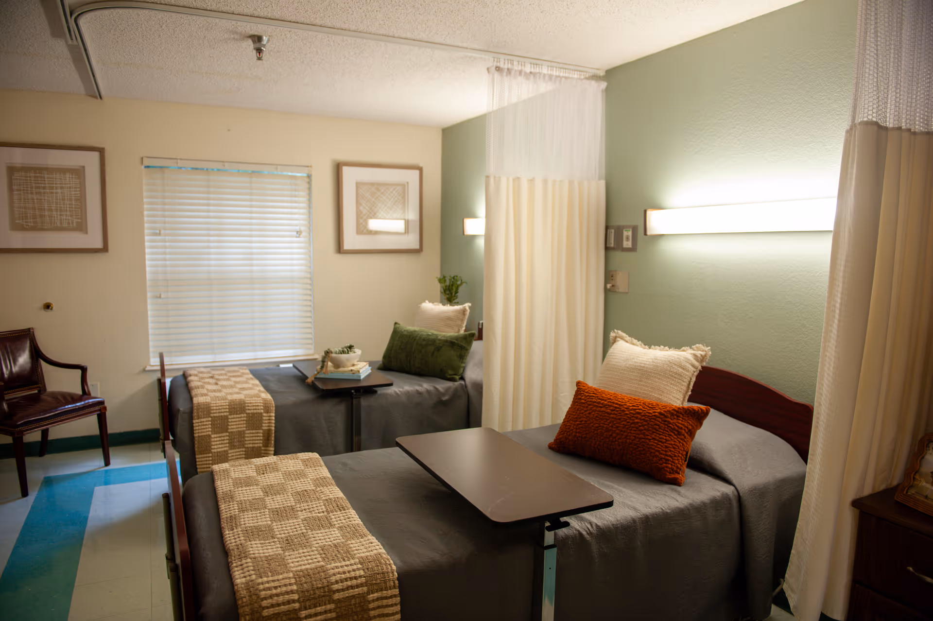 A cozy room with two single beds separated by a privacy curtain. Each bed has a gray bedspread, a beige checkered throw blanket, and decorative pillows in green, white, and orange. There is a small table on wheels positioned over each bed. The walls are painted light green and beige, with two framed artworks and a window with blinds. A wooden chair is placed near the window.