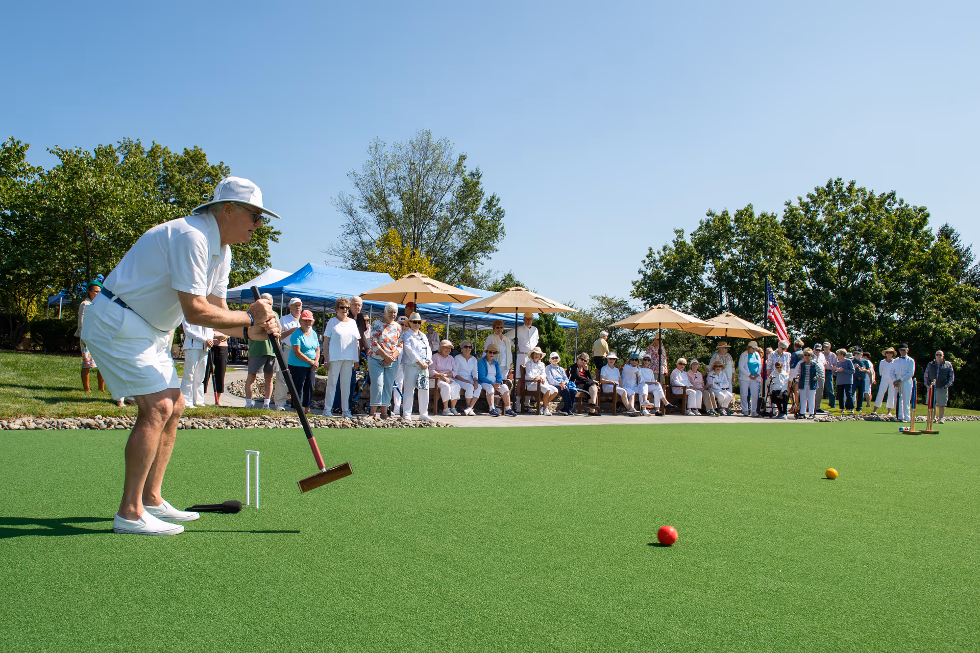 An elderly man in white clothing and a hat is playing croquet on a green lawn while a group of seniors watch from the sidelines under umbrellas and a blue canopy on a sunny day.