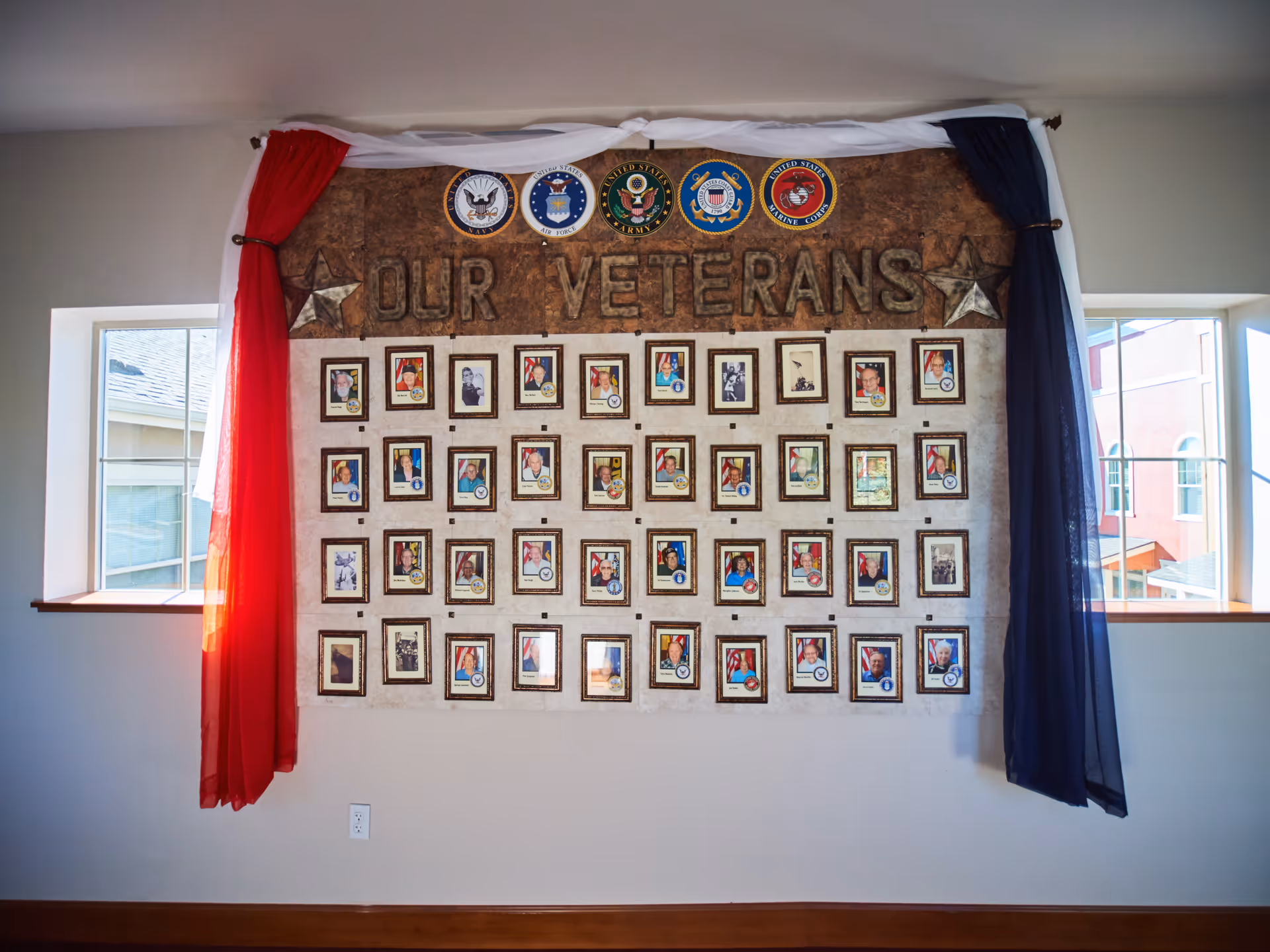 A wall display honoring veterans with framed photos of individuals arranged in rows. Above the photos are emblems of the United States Navy, Air Force, Army, Coast Guard, and Marine Corps, with the words 'OUR VETERANS' prominently displayed. The display is flanked by red, white, and blue curtains and is situated between two windows in a well-lit room.