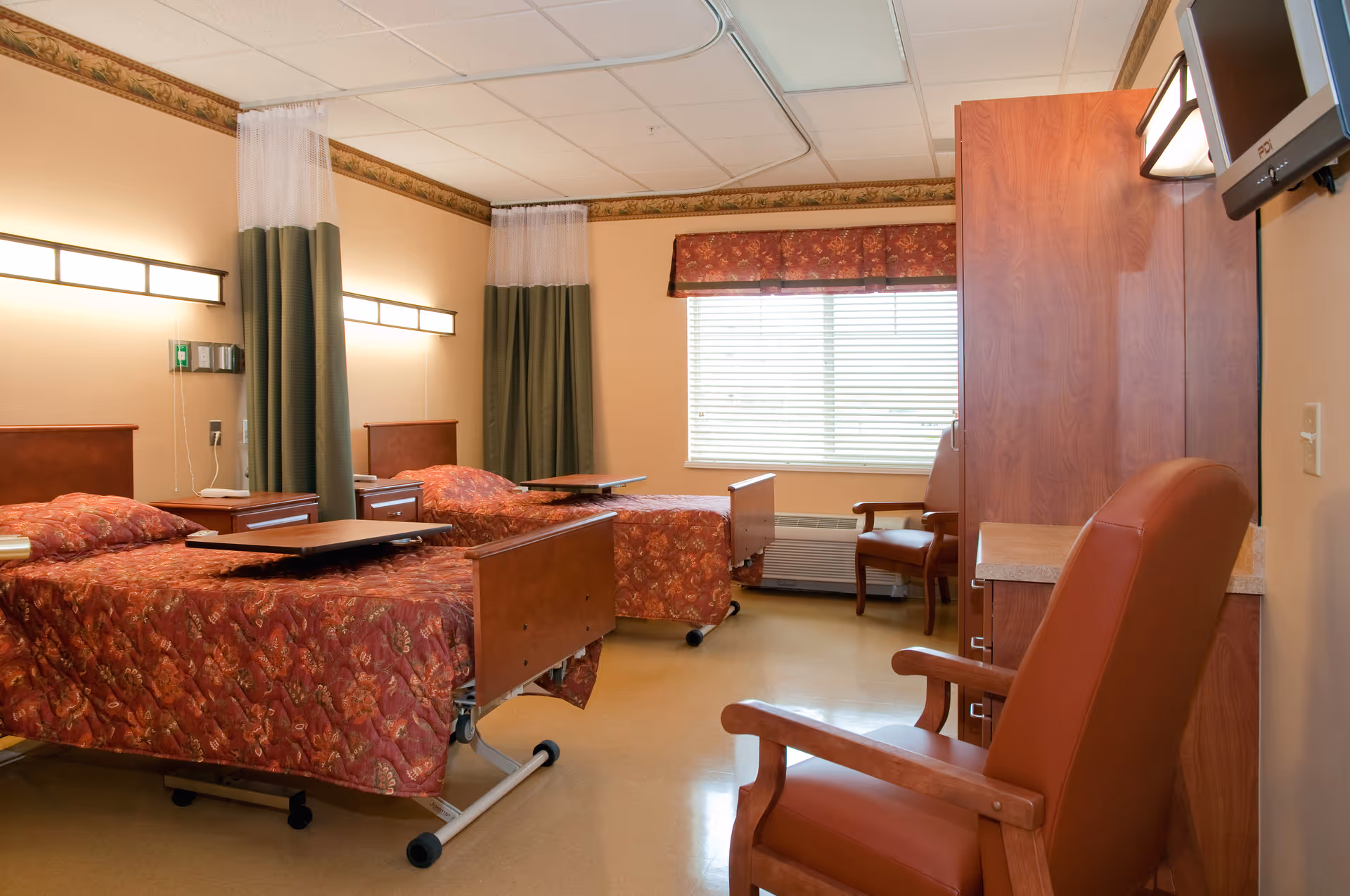 A room with two hospital-style beds covered with red patterned bedspreads, each with an overbed table. The room has beige walls, a window with blinds and a valance, two green privacy curtains, wooden furniture including a wardrobe and chairs, and a wall-mounted TV.