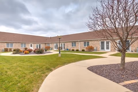 Outdoor courtyard area of a senior living facility with a paved walkway, green grass, landscaped bushes, a leafless tree, and a brick building with multiple windows and doors under a cloudy sky.