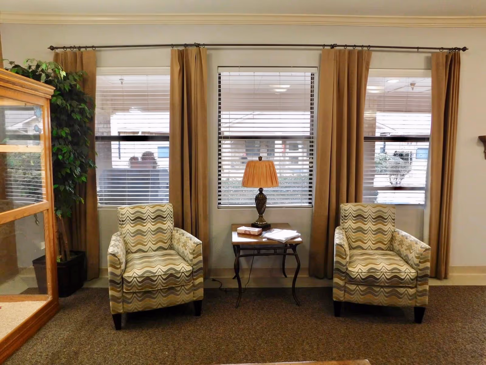 A cozy sitting area with two patterned armchairs separated by a small wooden table holding a decorative lamp and some books. Behind the chairs are three windows with beige curtains and white blinds. To the left, there is a tall green plant and a wooden display cabinet.