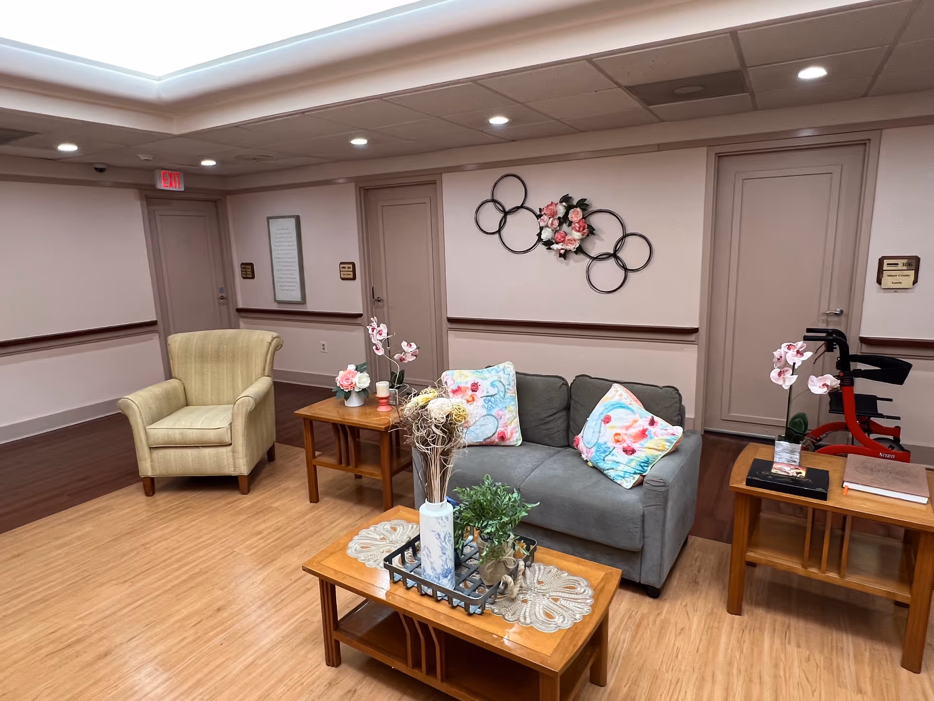 A cozy seating area in a senior living community featuring a gray couch with colorful floral pillows, a beige armchair, and three wooden tables adorned with decorative flowers and plants. The room has light-colored walls with a floral wall decoration and several closed doors in the background. The floor is a combination of wood and laminate, and a red walker is visible near one of the tables.