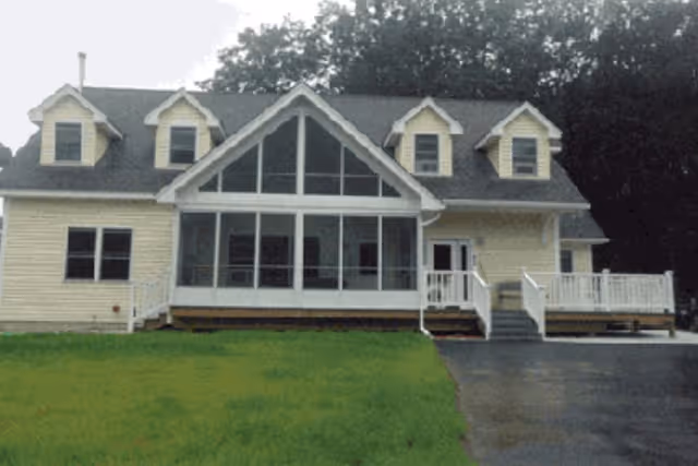 Front exterior view of a two-story house with a large central window section, three dormer windows on the roof, a white railing along the porch and steps, and a green lawn in front.