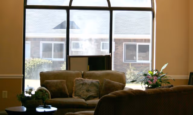 A cozy living room area with a large window showing the exterior of a brick building. The room has a beige couch with several pillows, a small round table with a plant, and another plant on the right side near the window.