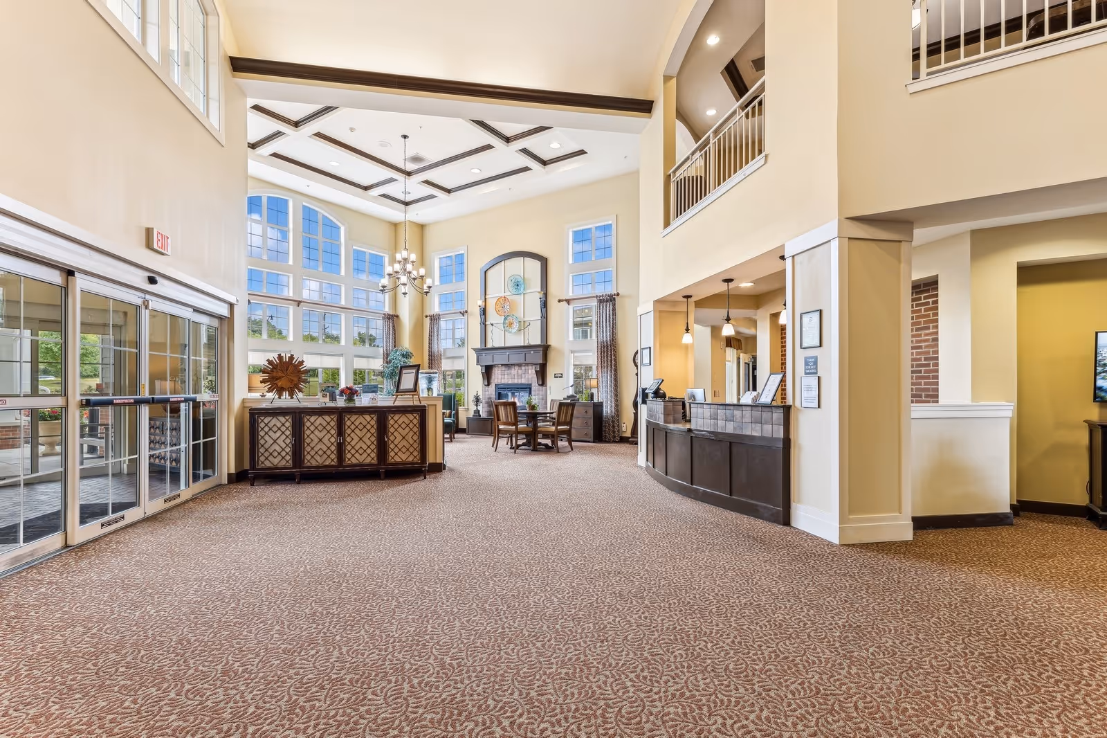 Spacious and well-lit lobby area of a senior living facility with large windows, high ceilings with decorative beams, a chandelier, a reception desk, and seating area near a fireplace. The entrance has automatic sliding glass doors.