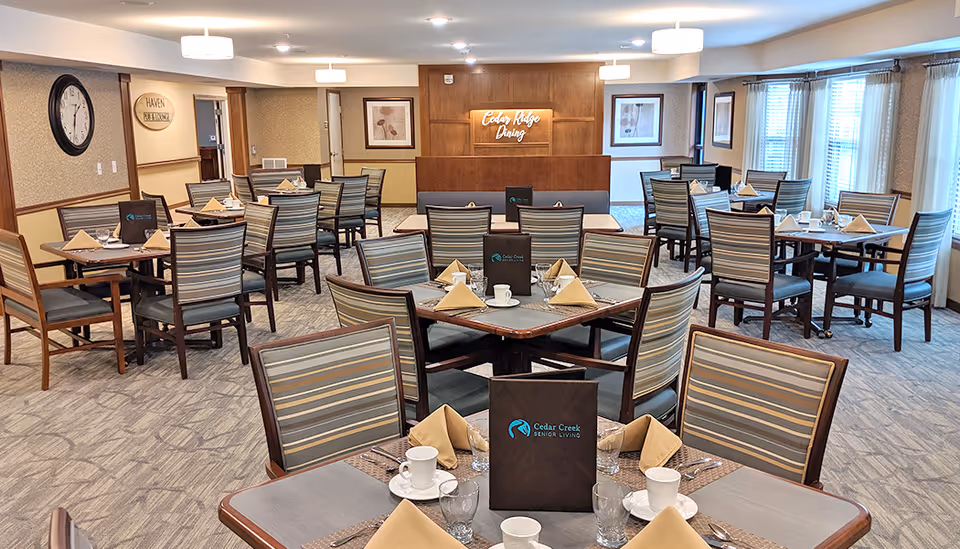 A spacious dining room in Cedar Creek Senior Living with multiple tables set with cups, glasses, napkins, and menus. The room has striped chairs, carpeted floors, framed artwork on the walls, and large windows with curtains allowing natural light. A sign on the back wall reads 'Cedar Ridge Dining'.