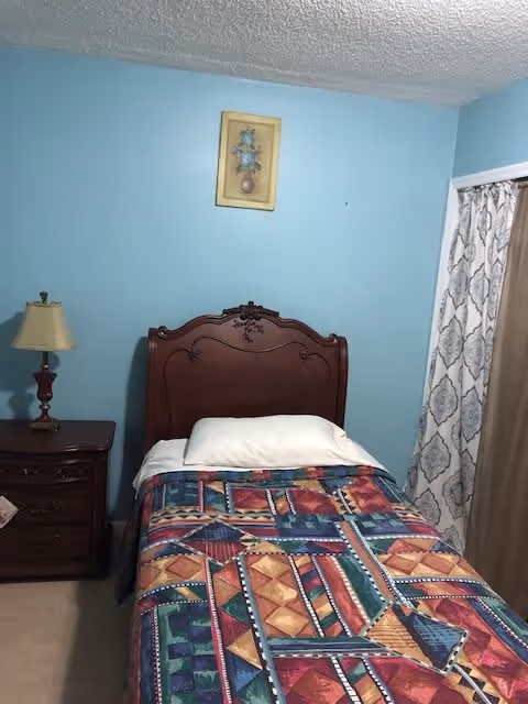 A small bedroom with a single bed featuring a colorful geometric patterned bedspread and a white pillow. The bed has a dark wooden headboard with carved details. To the left of the bed is a dark wooden nightstand with a lamp on it. The walls are painted light blue, and there is a framed picture hanging above the bed. On the right side, there is a window covered with patterned curtains.